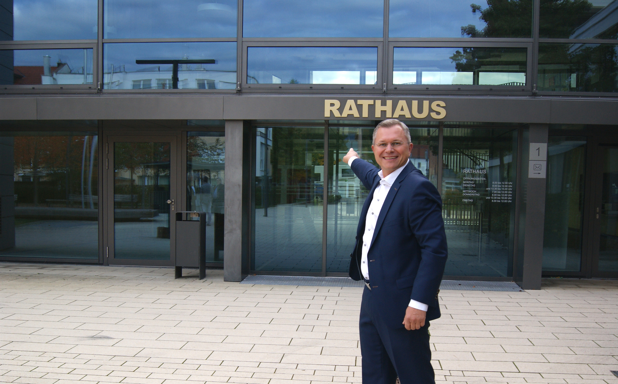 Stefan Siegl, kandidierender Bürgermeister der Gemeinde Gilching, steht im dunkelblauen Anzug vor dem Rathaus Gilching und zeigt lächelnd auf den Eingang.