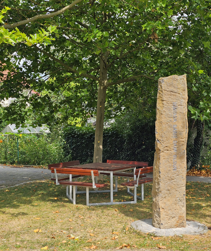 Spende Sitzgelegenheiten Gilching: Eine hölzerne Holzbank-Sitzgruppe im Schatten großer Bäume neben dem steinernen Friedenspfahl im Park.